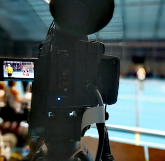 A camera on a tripod is prominently displayed with its viewfinder screen showing footage of an indoor sports event. The background is an indoor sports hall with a blue floor, and numerous spectators are faintly visible.