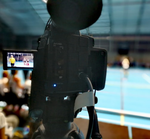 A camera on a tripod is prominently displayed with its viewfinder screen showing footage of an indoor sports event. The background is an indoor sports hall with a blue floor, and numerous spectators are faintly visible.