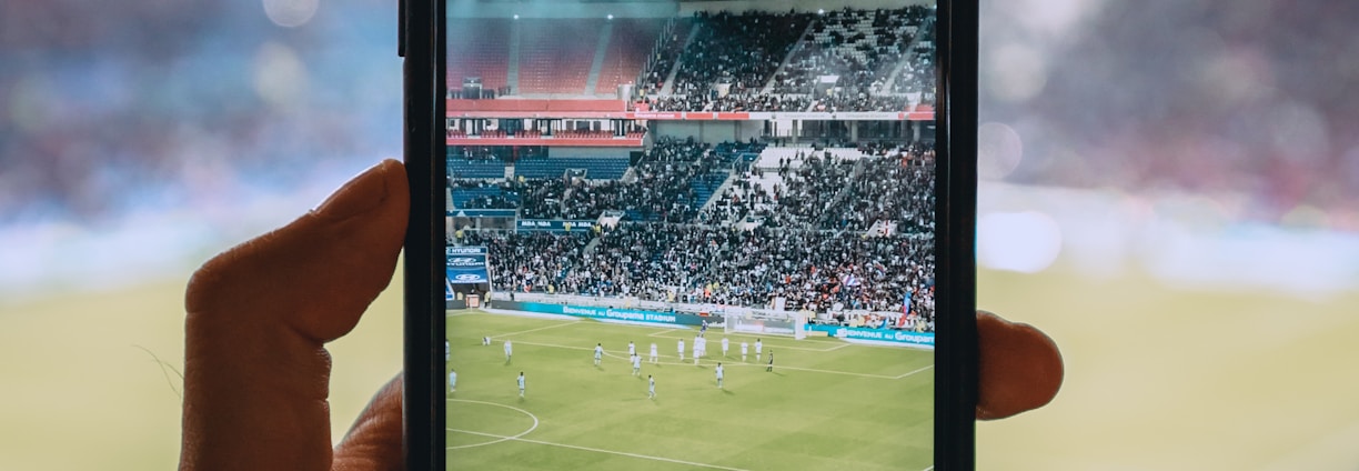 A hand holding a smartphone captures a live view of a soccer stadium filled with fans and players on the field. The image on the smartphone screen is crisp, showing a wide-angle view of the game in action.