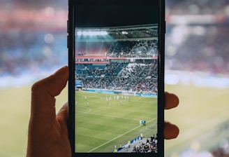 A hand holding a smartphone captures a live view of a soccer stadium filled with fans and players on the field. The image on the smartphone screen is crisp, showing a wide-angle view of the game in action.