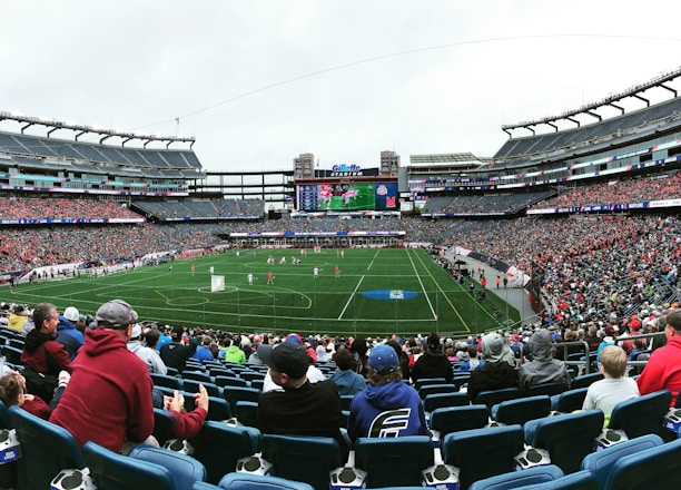 A large sports stadium filled with spectators. The field is green with white markings, and players in red and white uniforms are actively engaged in a game. The stands are packed with people wearing various colored clothing, and some sections of the crowd are more densely populated. Digital screens and advertisements are visible around the stadium, adding to the vibrant atmosphere.