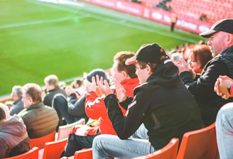 A group of people is sitting in bright red stadium seats, watching a live sports event. The stadium is well-lit with natural sunlight, and the grass on the field is vivid green. Most spectators are dressed in casual, warm clothing, reflecting cooler weather. The crowd is engaged, with many spectators clapping and attentive to the event.