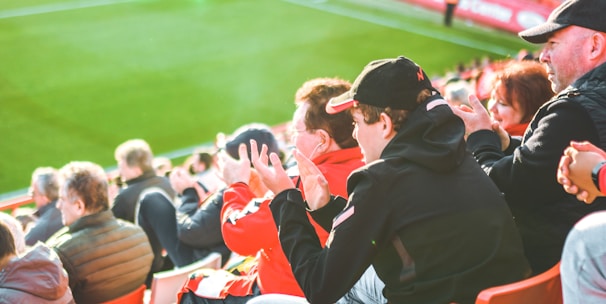 A group of people is sitting in bright red stadium seats, watching a live sports event. The stadium is well-lit with natural sunlight, and the grass on the field is vivid green. Most spectators are dressed in casual, warm clothing, reflecting cooler weather. The crowd is engaged, with many spectators clapping and attentive to the event.