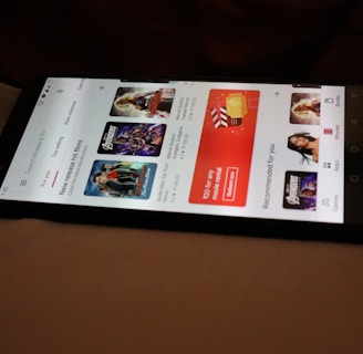 A smartphone displaying a streaming app interface, showing various movie and TV show thumbnails. The screen includes categories like 'For you' and 'New release hit films', with images from popular films. The background is a dimly lit setting with a fabric surface.