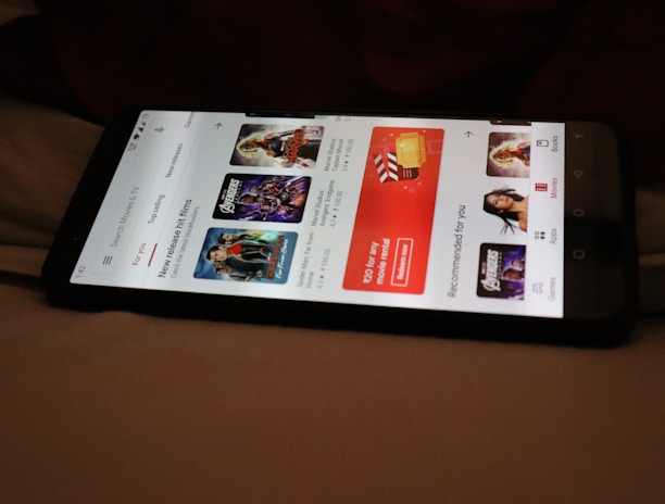 A smartphone displaying a streaming app interface, showing various movie and TV show thumbnails. The screen includes categories like 'For you' and 'New release hit films', with images from popular films. The background is a dimly lit setting with a fabric surface.