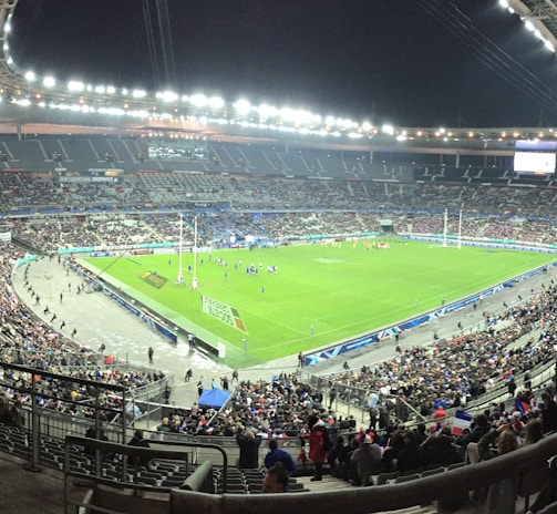 A large stadium filled with spectators, surrounding a green field where a sporting event is taking place. Bright stadium lights illuminate the area, with multiple banners and flags visible among the crowd. The architecture of the stadium is prominent, with tiered seating and large screens for viewing.