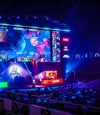 A large esports arena featuring a massive screen displaying a video game, surrounded by sponsor logos and team names. The auditorium is filled with spectators watching the gaming event. Neon blue and purple lights illuminate the stage, creating a vibrant atmosphere.
