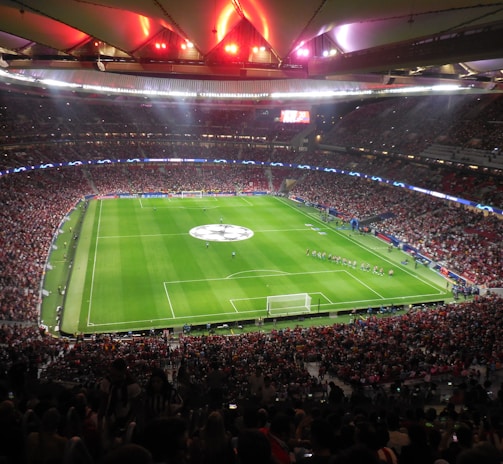 A large, packed stadium with bright lights illuminates a green soccer field. The scene is vibrant with a massive audience surrounding the pitch, visibly excited and engaged. The center of the field displays a prominent circular logo, suggesting a major sporting event. Players and staff can be seen on the field, likely preparing for a match.