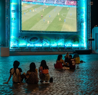 A large outdoor screen displays a soccer match at night. A small group of people sit on a cobblestone surface, watching the game. The screen is surrounded by a bright blue glow, creating a lively atmosphere.