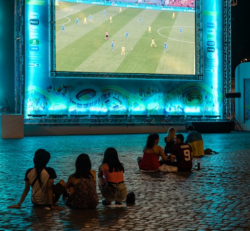 A large outdoor screen displays a soccer match at night. A small group of people sit on a cobblestone surface, watching the game. The screen is surrounded by a bright blue glow, creating a lively atmosphere.