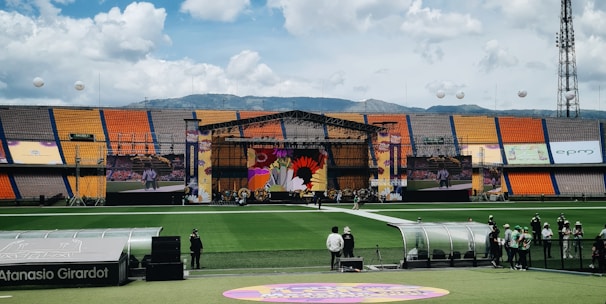 A sports stadium with a large stage set up on the field, hosting an outdoor event. Spectators wearing colorful hats sit in the foreground. The seating areas display vibrant colors, and a clear blue sky with scattered clouds is visible. Large screens display graphics related to the event.
