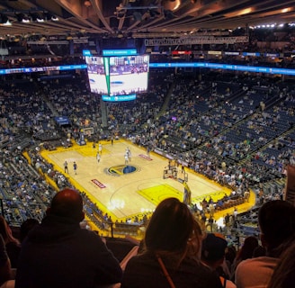 An indoor basketball arena filled with spectators. The court features a central logo and is surrounded by partially occupied tiered seating. A large scoreboard hangs above the center of the court, displaying the game score and time. Ambient lighting highlights the players on the court and the vibrant atmosphere of a live sports event.
