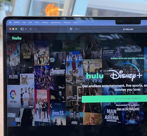 A laptop screen displays a web page featuring a mix of movie and TV show covers, highlighting popular titles and a Hulu and Disney+ bundle promotion. There are visible icons and images of films and shows alongside promotional text offering entertainment and live sports.