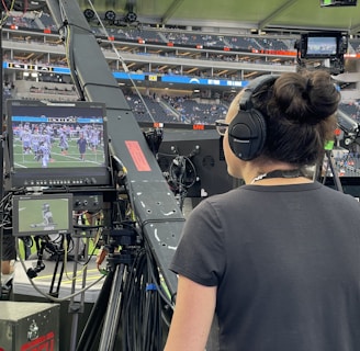 A person with headphones is operating a large camera setup at a sports stadium. The camera equipment is focused on a screen showing a live sports event taking place on a football field. The stadium is filled with spectators, and the atmosphere is busy with technical setup and monitors.