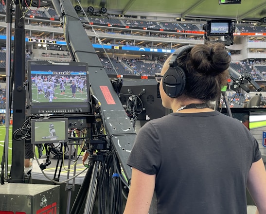 A person with headphones is operating a large camera setup at a sports stadium. The camera equipment is focused on a screen showing a live sports event taking place on a football field. The stadium is filled with spectators, and the atmosphere is busy with technical setup and monitors.