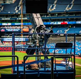 A professional-grade video camera is mounted on a platform overlooking a baseball field. The stadium seats in the background are filled with some spectators, and various advertisements are visible around the field. A staff member wearing a jacket with 'STAFF' written on the back is positioned near the camera.