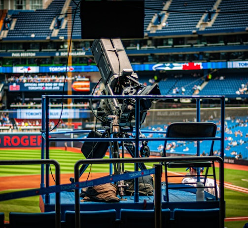 A professional-grade video camera is mounted on a platform overlooking a baseball field. The stadium seats in the background are filled with some spectators, and various advertisements are visible around the field. A staff member wearing a jacket with 'STAFF' written on the back is positioned near the camera.