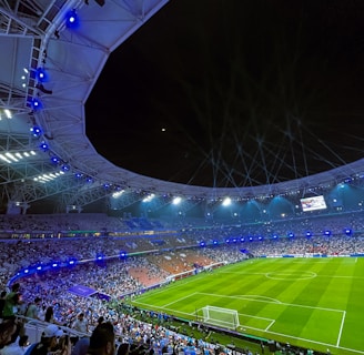 A large, illuminated stadium filled with spectators, with a focus on a well-maintained football field and energetic crowd. Bright stadium lights cast a glow over the scene, enhancing the lively atmosphere as people watch an ongoing sporting event.
