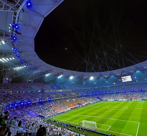 A large, illuminated stadium filled with spectators, with a focus on a well-maintained football field and energetic crowd. Bright stadium lights cast a glow over the scene, enhancing the lively atmosphere as people watch an ongoing sporting event.